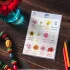 A chart of flowers name in Punjabi is on the wooden table with a flower in a glass container, colored pencils, and a colorful bead necklace around it.