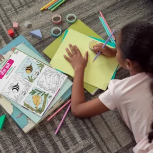 A coloring book is placed on other books near a child drawing on a chart, with tapes, pencils, and erasers on the carpet.