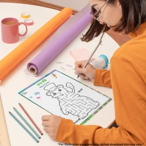 A girl is drawing in a difficult maze puzzle book on the table. There are pencils, chart papers, notes, cello tapes, a ruler, and a cup around it.