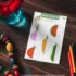 A trace and color book is placed on a wooden table with a flower in a glass container, some crayons, and a colorful beads necklace beside it.