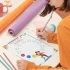 A girl wearing orange clothes is writing in a coloring page pattern book on the table, with pencils, chart papers, tapes, a sharpener, and a cup around.