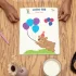 Two hands show a preschool tracing sheet on the wooden table, with a glass of milk, a black plate, decorative items in a container, and a pencil nearby.