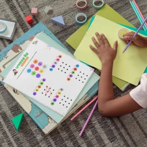 Preschool activity pages are placed on other books near a child drawing on chart paper, with pencils, erasers, and cello tapes on the floor.